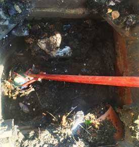 Zoomed view of a blocked drain half-full of old stagnant water, showing a build-up of mainly vegetation and litter. A specialist is clearing the blockage using a long red grabber rod.