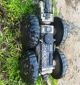 Shiny all-metal CCTV survey vehicle with large thin black rubber whhels lightly coated in fresh mud after tracing and mapping a drainage system