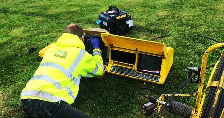 Cctv survey specialist dressed in bright yellow high visibility clothing laying on a grass bank looking at a monitor enclosed in a yellow plastic casing dispaying an underground pipe's internal structure which is mostly obscured by the specialists hand