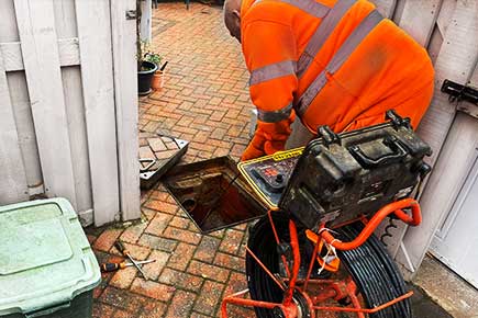 CCTV drain-surveying technician in orange high-visibility clothing feeding a black cable from a circular metal reel into a drain beneath a red brick driveway