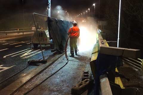 A contract Drains Pipes Solutions specialist operative, wearing orange upper and yellow lower workwear plus a white hard hat, using high-pressure jetting equipment to thoroughly clean a motorway drainage system.