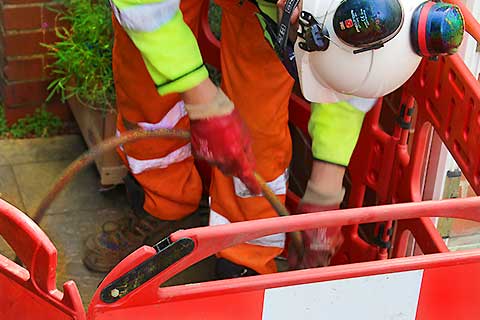 Close-up photo of a jetter wearing a fluorescent yellow jacket and fluorescent orange trousers, using jetting equipment and holding a pipe that disappears down a kitchen sewer drain.