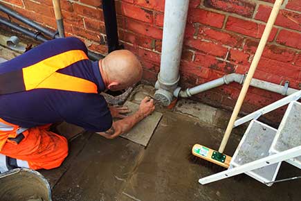 A kitchen sink pipe at the side of a house disappearing below ground; a specialist kneels beside it, laying the final new paving after excavating, clearing and repairing the drain.