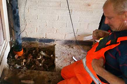 Drain repair expert in orange high-visibility overalls kneeling at an excavated kitchen floor, working on a sewer repair that continues further underground