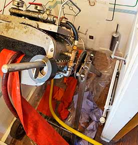 Zoomed-in top view of a sluice-liner assembly with a large oval metal body secured by multiple bolts, pipes, and fittings, used to reline a damaged sewer pipe connected to a home�s downstairs loo