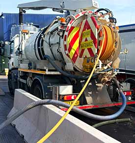 Distant rear-corner view of a large white drainage-lining vehicle with fluorescent orange and yellow diagonal chevron stripes on the back panel, and two pipes extending from the vehicle down into a Sheffield drain