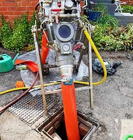 Front-facing close-up of a sluice-lining machine with a polished metal body and framework, featuring gauges for pump and air pressures, with a slim yellow pipe and a thicker orange pipe running down into exterior residential kitchen drains
