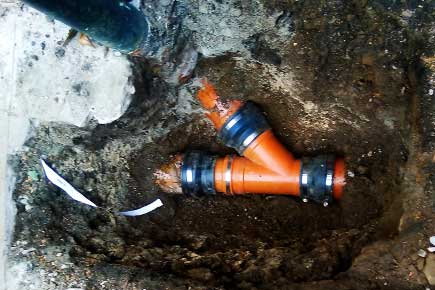 Close-up of a three-junction underground drainage repair using a new mild-brown junction pipe installed within an excavated channel