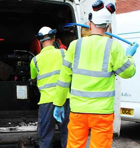 Two drain and sewer lining engineers in high-visibility clothing with hard hats and noise-cancelling ear protection standing behind a white van with its rear doors open, unloading pipe-repairing equipment