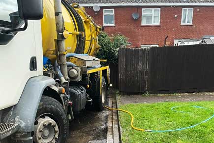 Front-right three-quarter view of a bright yellow drain truck facing the camera, parked beside a grass verge outside a brick-built residential property in Sheffield.