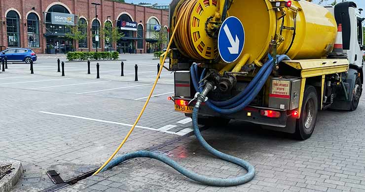 Distance rear view of our bright yellow unmarked jet-vac vehicle, with a pipe feeding into the drains at a Sheffield commercial retail car park, clearing and removing built-up drainage debris.