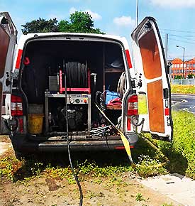Rear view of one of our white company vehicles, parked on a roadside verge with both rear doors wide open. Drain and sewer jetting equipment is visible inside, with a black hose disappearing underground.